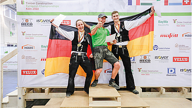 Drei Männer auf Siegertreppchen aus Holz. Männer links und rechts in Zimmermannskleidung und Deutschlandfahne in der Hand.