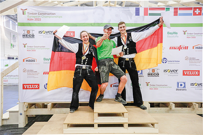 Die Gewinner v. l. n. r.: Benedikt Pfister (Bronze), Marcel Bolego (Gold) und Philipp Kaiser (Silber) Drei Männer auf Siegertreppchen aus Holz. Männer links und rechts in Zimmermannskleidung und Deutschlandfahne in der Hand.