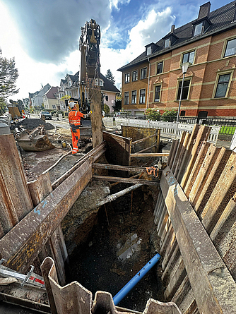 Verbauter Graben einer Kanalbaustelle mit Bauarbeiter und Bagger auf einer Straße.