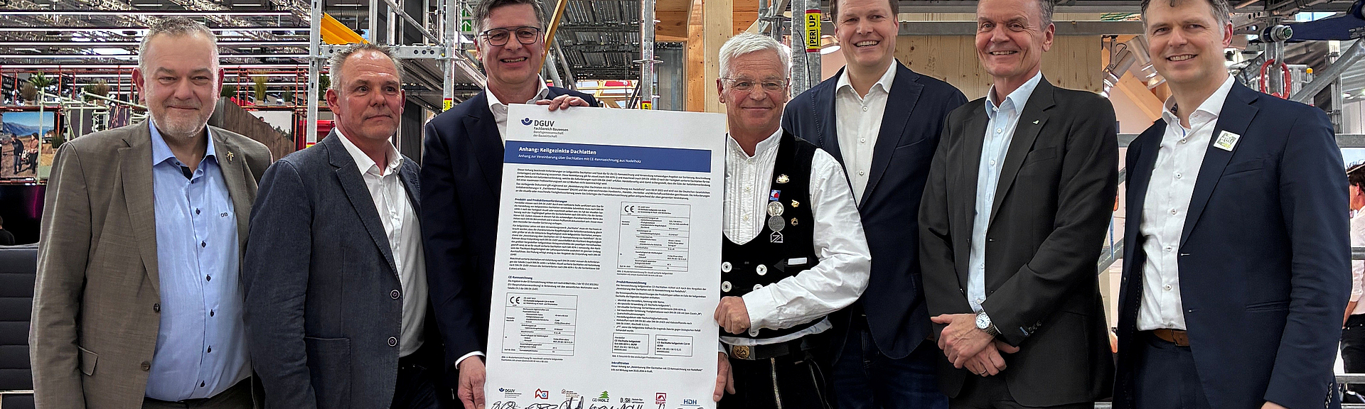 Sieben Männer stehen auf einem Messestand der BG BAU zusammen und präsentieren ein unterzeichnetes Dokument zur Dachlattenvereinbarung vor einer Gerüstkonstruktion und einer großen Bildschirmanzeige.
