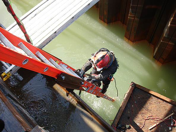 Trübes Wasser, schlechte Sicht, beengte Verhältnisse: Das professionelle Tauchen im Wasser an Baustellen ist eher kein Freizeitvergnügen. Ein Taucher klettert eine Leiter herunter zu einem Kanal, der mit Wasser gefüllt ist.