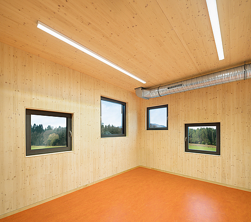 Innenraum mit Holzwänden und vier Fenstern in unterschiedlicher Höhe und Größe. An der Decke führt ein Rohr an.