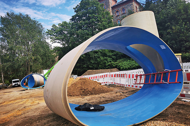 Aufgrund ihrer geringeren Wanddicke ist bei den GFK-Rohren eine größere Überdeckung möglich als bei Stahlbetonrohren desselben Profils. Großaufnahme des ovalen GFK-Rohres. Eine Leiter, die fast so lang wie das Rohrstück ist, und eine Jacke im Rohrstück verdeutlichen die Dimension.