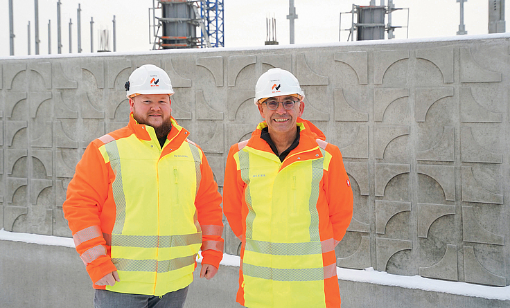 Der Projektleiter und der Bauleiter stehen mit Helm und gelb-oranger Jacke vor einer Betonwand der Schule.