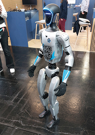 Humanoider Roboter mit schwarzem Visier und leuchtendem Rahmen und QR-Code läuft über die Messe digitalBAU 2026 in Köln. 