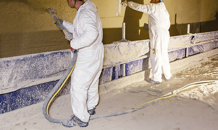 Das Team arbeitet an einer Tunnelwand: Zwei Beschäftigte tragen Schutzanzüge, Arbeitshandschuhe und Sicherheitsschuhe beim Aufspritzen und Glätten des Brandschutzputzes.