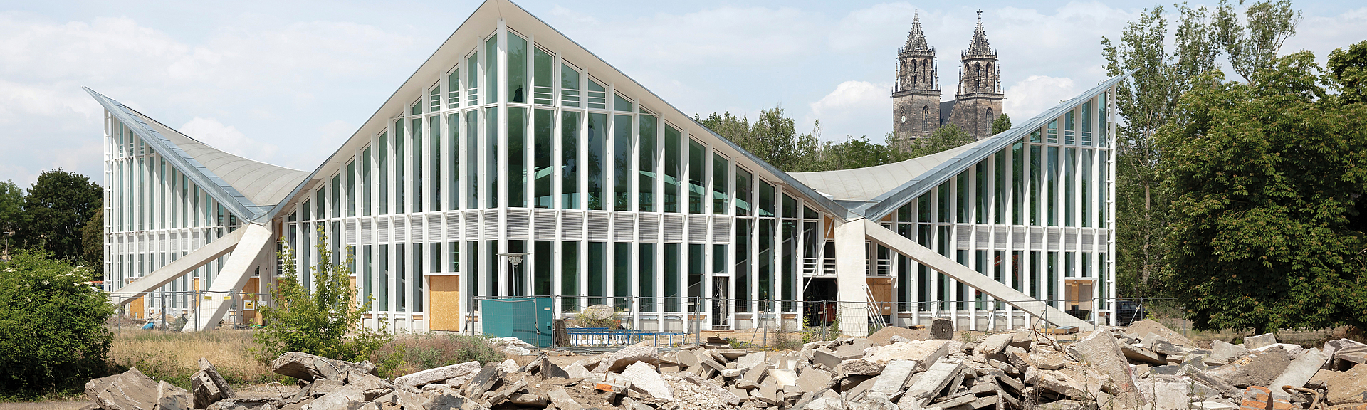 Hyparschale Magdeburg: Modernes Gebäude mit gefalteter Glas-Stahl-Dachkonstruktion; davor Bauschutt auf einer Baustelle.
