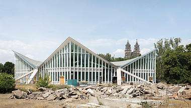Hyparschale Magdeburg: Modernes Gebäude mit gefalteter Glas-Stahl-Dachkonstruktion; davor Bauschutt auf einer Baustelle.