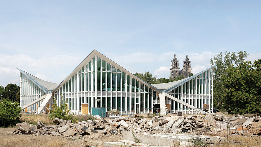 Hyparschale Magdeburg: Modernes Gebäude mit gefalteter Glas-Stahl-Dachkonstruktion; davor Bauschutt auf einer Baustelle.