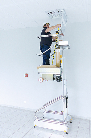 Ein Mann mit Schutzhelm steht auf einer fahrbaren Ein-Person-Hubarbeitsbühne und führt Arbeiten an der Deckenbeleuchtung durch.