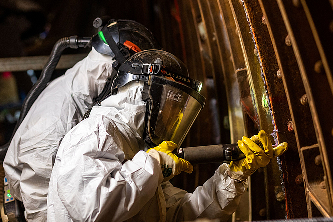 Zwei Personen in Vollschutzkleidung (gebläseunterstützter Helm mit Partikelfilter und Schutzanzug Kategorie III Typ 5 sowie Nitrilhandschuhe) beim Verstemmen der Bleifugen im Tunnelinneren.