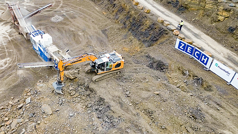 Vogelperspektive auf Bagger, der eine Brechanlage belädt.