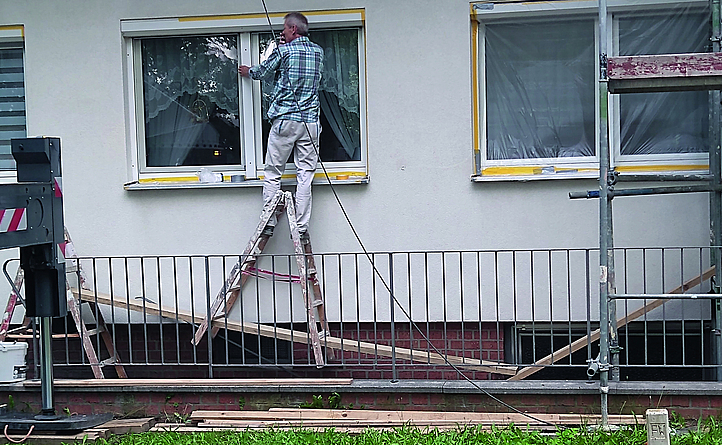 Negtivbeispiel - Ein Maler steht auf den obersten Stufen einer Holzleiter. Diese steht auf einer selbstgebauten Leiter- und Bretterkonstruktion, die jederzeit verrutschen kann. 