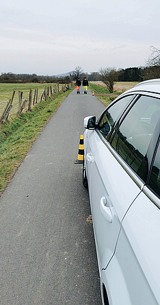 Im Hintergrund stehen zwei Personen auf einer kleinen Landstraße. Im Vordergrund ist ein Auto, vor dem ein Warnkegel steht. Die Person rechts hat gelbe Hosen an, die Person links orange-rote.