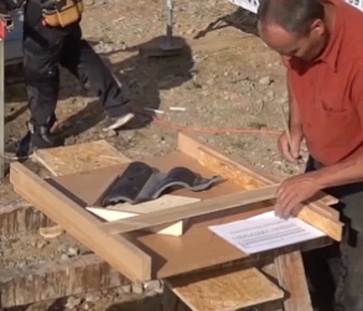 Ein Mann steht mit Stift in der Hand vor einer Schablone, die sich auf einem Holzgestell befindet. In der Schablone liegen ein Ziegel und eine Liste.