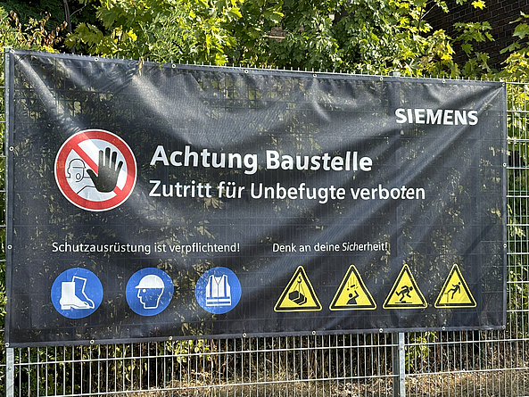 Auf der Plane mit Siemens-Logo und Piktogrammen wird auf die Pflicht zur Nutzung von Sicherheitsschuhen, Helm und Warnweste hingewiesen sowie vor schwebenden Lasten, herabfallenden Gegenständen, Stolperstellen und Absturzgefahr gewarnt.
