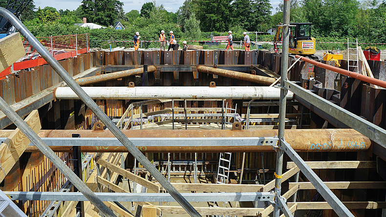 Blick in einen mit Stahlträgern ausgesteiften Spundwandkasten auf einer Baustelle; mehrere Personen stehen am Rand der Baugrube.