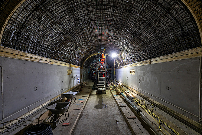 in einem beleuchtenden Tunnel steht eine Person in oranger Warnkleidung auf einer Leiter und bearbeitet die Decke.