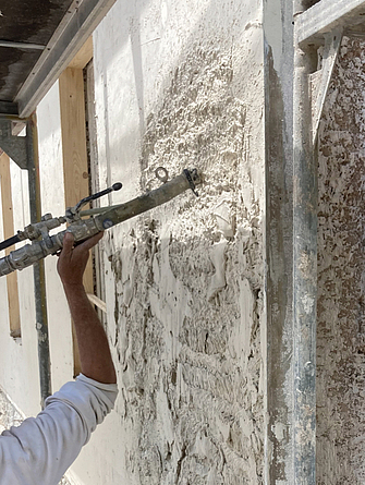 Eine Person trägt mit einer Spritze helles Material auf eine Fassade auf.