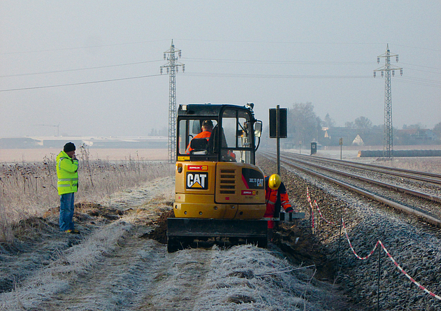 Einsatz einer rot-weißen-Kette ohne Sicherungsmaßnahmen gegen Gefahren aus dem Bahnbetrieb direkt an der Grenze des Gleisbereichs In und um einen Bagger arbeiten Gleisarbeiter direkt an der Abgrenzung mit einer rot-weißen Kette.