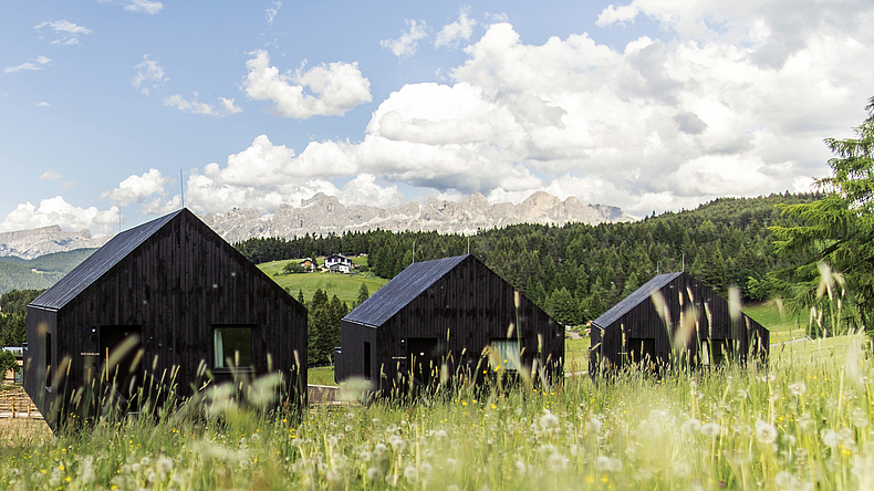 Drei kleine, schwarze Holzhäuser mit Giebeldach in bergiger, grüner Landschaft.
