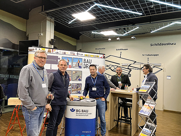 Der Informationsstand der BG BAU mit großer bedruckter Leinwand im Hintergrund und Messetisch, an dem mehrere Männer stehen, im Vordergrund.