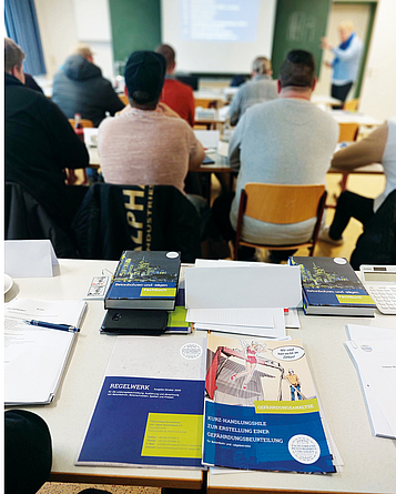 Teilnehmer und Referent im Seminarraum von hinten gesehen. Im Vordergrund ausgebreitetes Regelwerk auf dem Tisch.