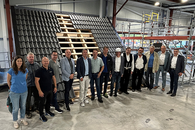 Eine Gruppe von Personen steht in einer Werkhalle vor einem großen, teilweise gedeckten Dachmodell.