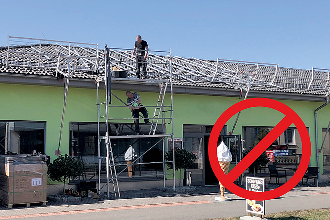 Haus, an dem teilweise Seitenschutzsysteme angebracht sind. Auf dem Dach steht ein ungesicherter Mann und blickt auf einen anderen Mann der auf der ersten Gerüstebene an der Vorderseite des Hauses steht.