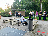 Ein Mann in weißem Kittel führt knieend auf einem gepflasterten Platz ein Experiment durch. Mehrere Personen beobachten ihn aus sicherer Entfernung 