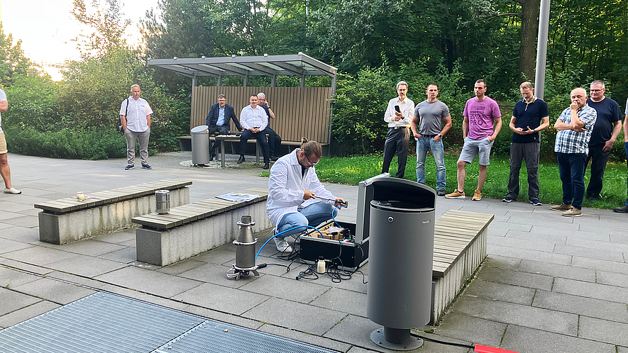 Demonstration von Staubexplosionen Ein Mann in weißem Kittel führt knieend auf einem gepflasterten Platz ein Experiment durch. Mehrere Personen beobachten ihn aus sicherer Entfernung