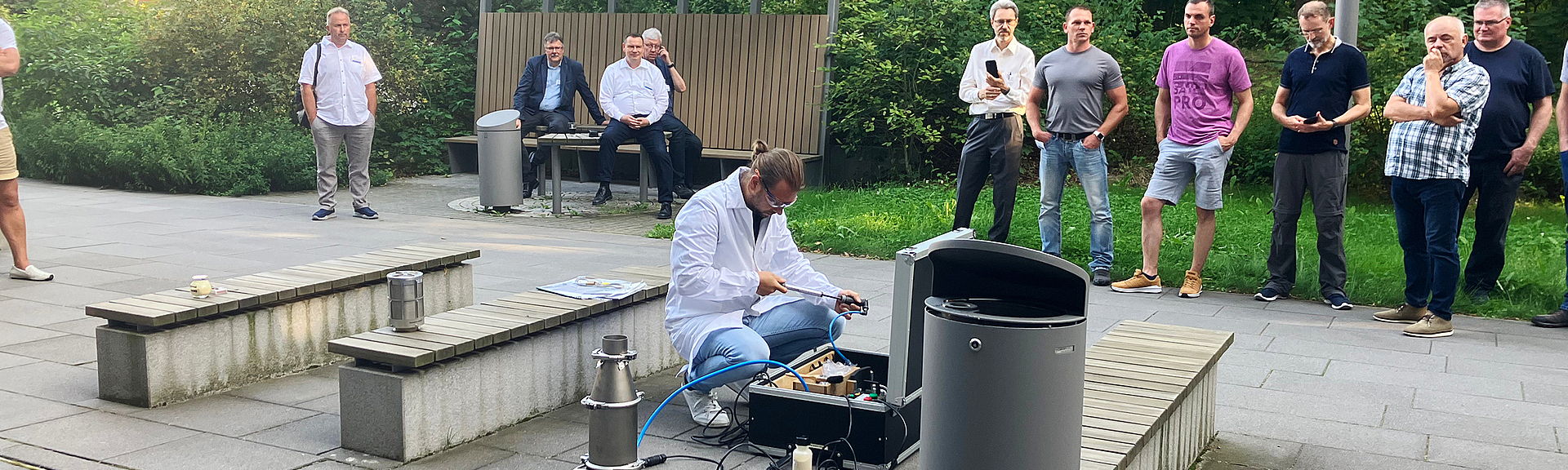 Ein Mann in weißem Kittel führt knieend auf einem gepflasterten Platz ein Experiment durch. Mehrere Personen beobachten ihn aus sicherer Entfernung 