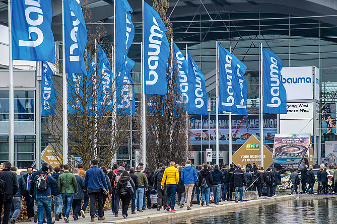 Menschenmassen bewegen sich auf den Eingang der bauma-Messehallen zu und passieren Fahnenmasten mit bauma-Logo.