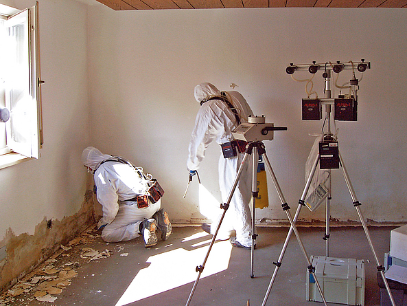 Zwei Personen in Schutzanzügen arbeiten in einem Raum mit beschädigter Wand. Sie entfernen Material nahe dem Boden. Neben ihnen steht ein Messgerät auf einem Stativ zur Erfassung von Schadstoffen.