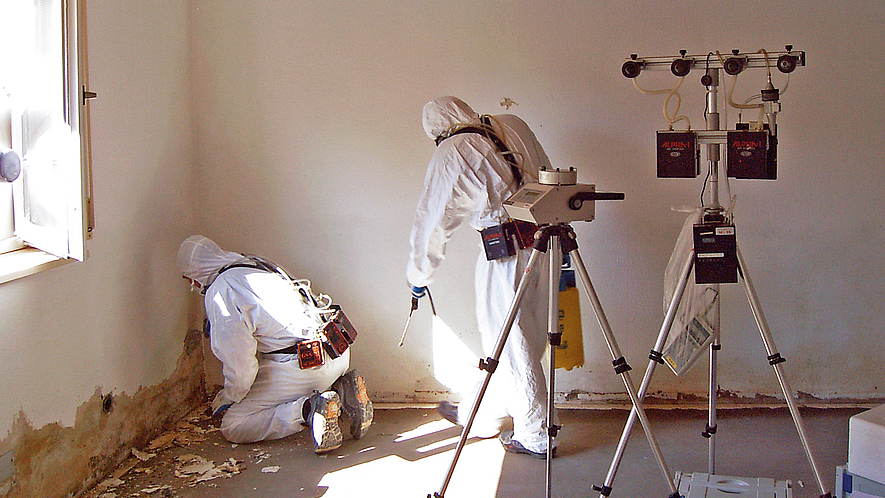 Arbeitsplatzmessung biologischer Arbeitsstoffe Zwei Personen in Schutzanzügen arbeiten in einem Raum mit beschädigter Wand. Sie entfernen Material nahe dem Boden. Neben ihnen steht ein Messgerät auf einem Stativ zur Erfassung von Schadstoffen.