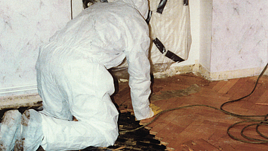 Eine Person in weißem Schutzanzug kniet in einem Raum und entfernt beschädigte Bodenbeläge. Teile des Bodens sind freigelegt.