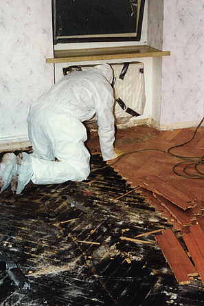 Eine Person in weißem Schutzanzug kniet in einem Raum und entfernt beschädigte Bodenbeläge. Teile des Bodens sind freigelegt.