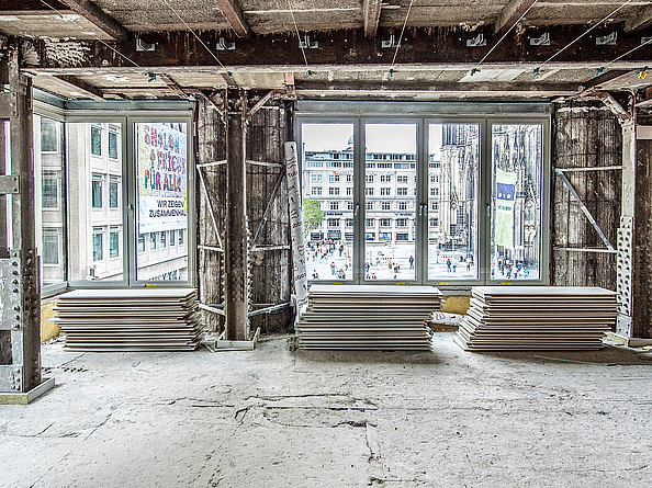 Baustelle in einem Gebäude, in dem Decken- und Fußbodenarbeiten durchgeführt werden. An der Fensterfront liegen übereinandergestapelte Bodenplatten.