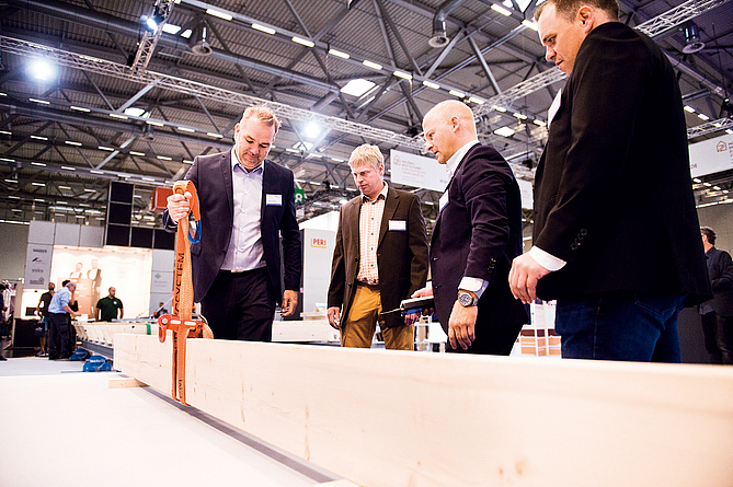 Zwei Männer schauen an einem Messetisch zu, wie ein dritter ein Holzstück an einem Seil mit Automatikhaken hochzieht.