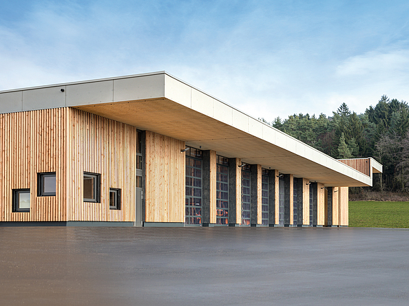 Seitenansicht des Bauhof-Gebäudes mit Holzfassade und großen Glastoren auf betonierter Fläche im Vorfeld.