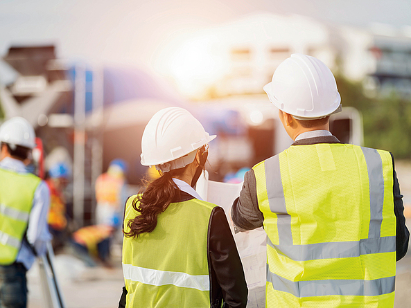 Eine Frau und ein Mann, in Schutzhelm und Warnweste, schauen gemeinsam auf eine Planskizze. Sie befinden sich auf einer Baustelle.