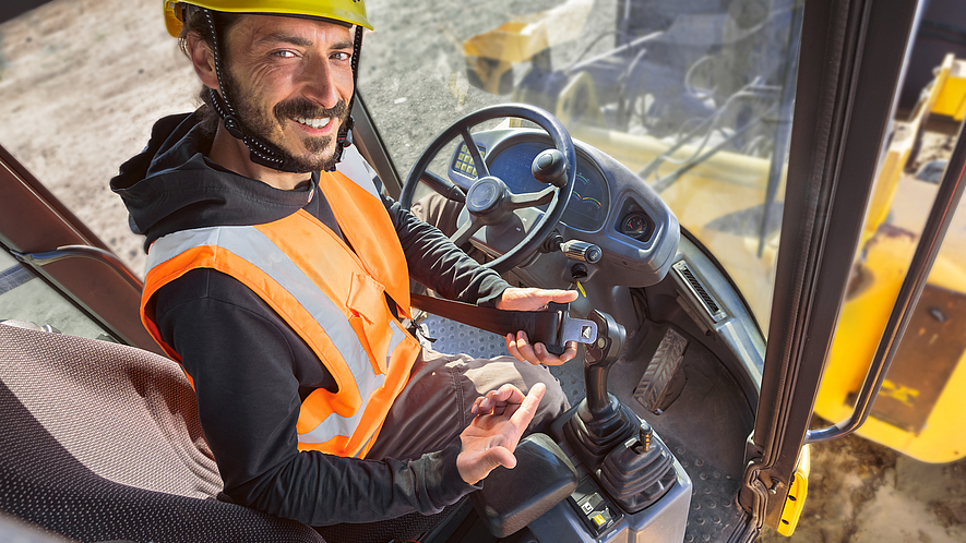 Motiv Anschnallkampagne Ein Bauarbeiter deutet mit dem Finger auf den Sicherheitsheitgurt. Er sitzt in einer Baumaschine.