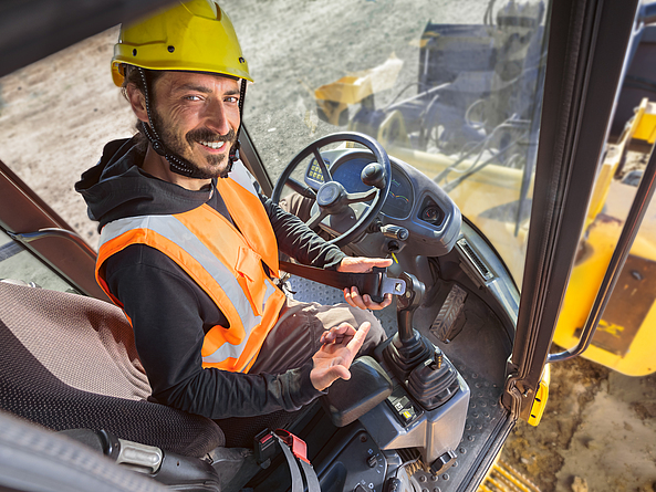 Ein Bauarbeiter deutet mit dem Finger auf den Sicherheitsheitgurt. Er sitzt in einer Baumaschine.