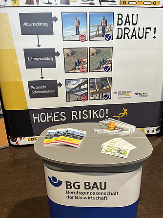 Hinter einem Ausstellungstisch mit ausgelegten Prospekten steht ein Roll-up der BG BAU mit lebenswichtigen Regeln.