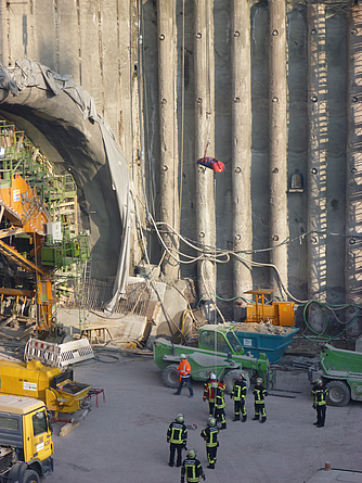 Baustelle mit Tunnelöffnung, Maschinen und Einsatzkräften; eine waagerechte Trage hängt gesichert an einem Seil in der Luft.