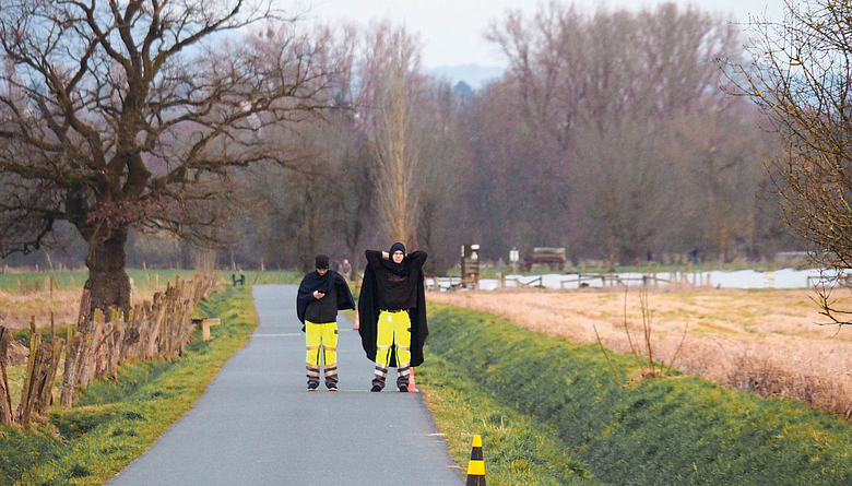 Zwei Personen mit gelben Warnhosen, schwarzer Oberbekleidung und schwarzen Mützen stehen auf einer kleinen Landstraße. 