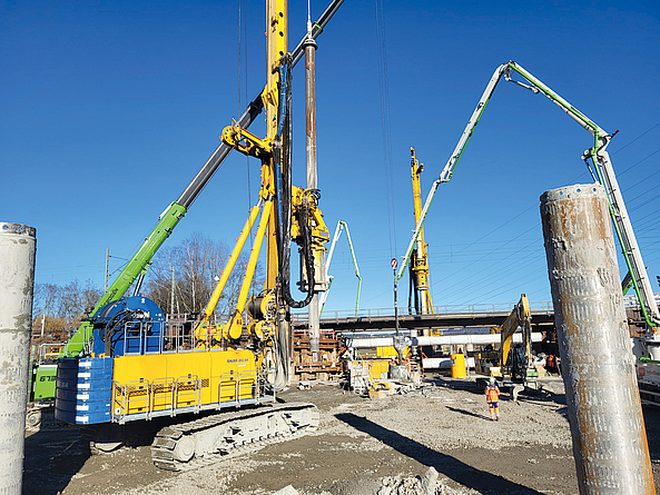 Bei den Bohrarbeiten kommen u. a. die Großdrehbohrgeräte BAUER BG 39 und BAUER BG 46 zum Einsatz. Baustelle mit verschiedenen Bohrgeräten in Aktion, im Hintergrund ist die Eisenbahnstrecke zu sehen.