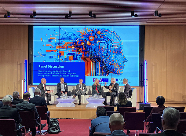 Sechs Personen sitzen auf einer Bühne und diskutieren im Rahmen einer Paneldiskussion vor einem großen Bildschirm. Auf dem Bildschirm steht der Titel der Diskussion über Chancen transnationaler KI-Lösungen für Sicherheit und Gesundheit bei der Arbeit.