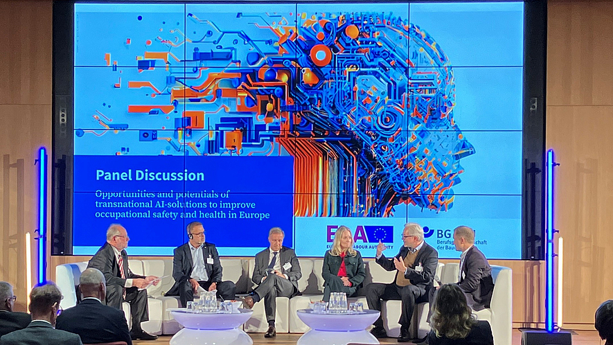 Sechs Personen sitzen auf einer Bühne und diskutieren im Rahmen einer Paneldiskussion vor einem großen Bildschirm. Auf dem Bildschirm steht der Titel der Diskussion über Chancen transnationaler KI-Lösungen für Sicherheit und Gesundheit bei der Arbeit.
