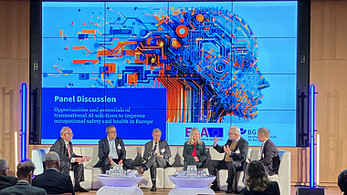 Sechs Personen sitzen auf einer Bühne und diskutieren im Rahmen einer Paneldiskussion vor einem großen Bildschirm. Auf dem Bildschirm steht der Titel der Diskussion über Chancen transnationaler KI-Lösungen für Sicherheit und Gesundheit bei der Arbeit.